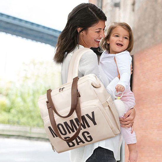 Maman souriante porte sa petite fille dans les bras avec un sac a langer bebe sur l’epaule